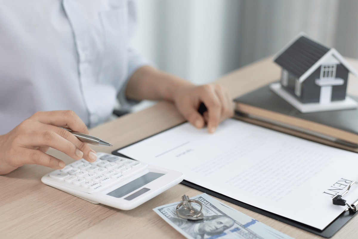 A woman signs a contract, calculating figures with a house model nearby, ensuring a secure transaction.