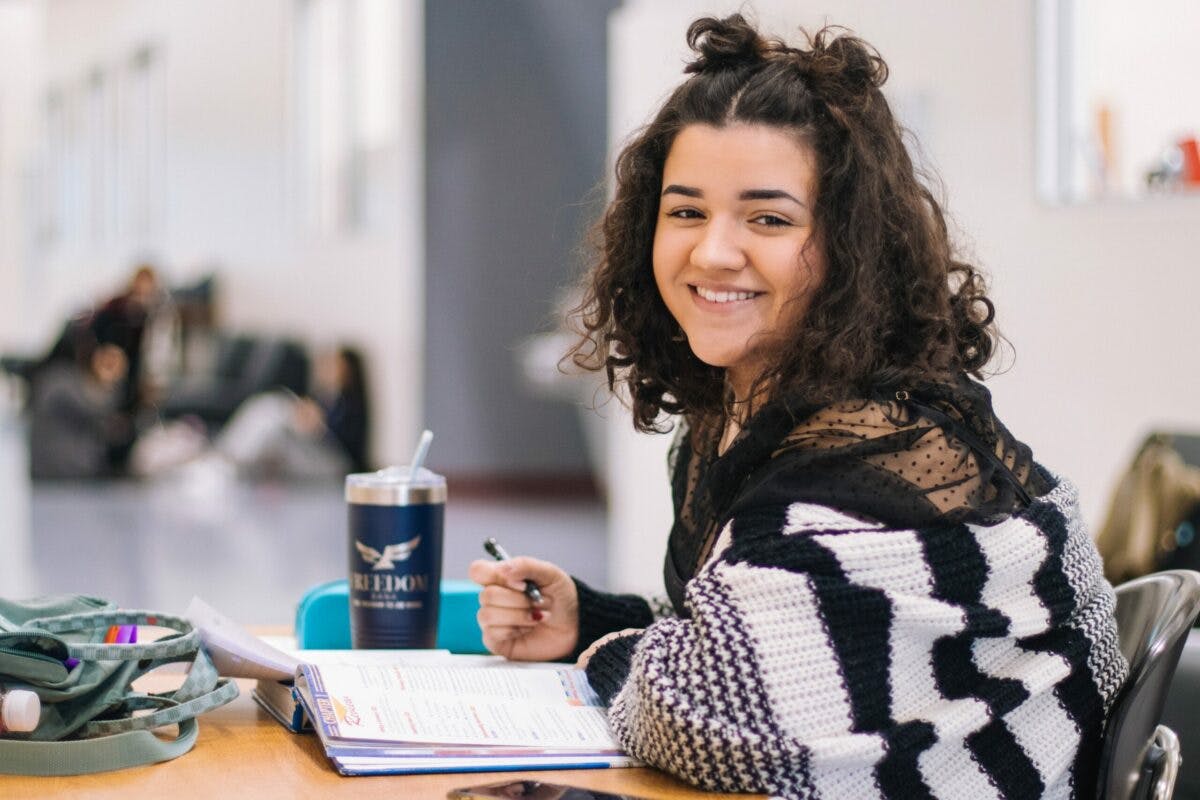 A student, wearing a smile, sits at a table, engrossed in her notebook, showcasing her dedication to learning.