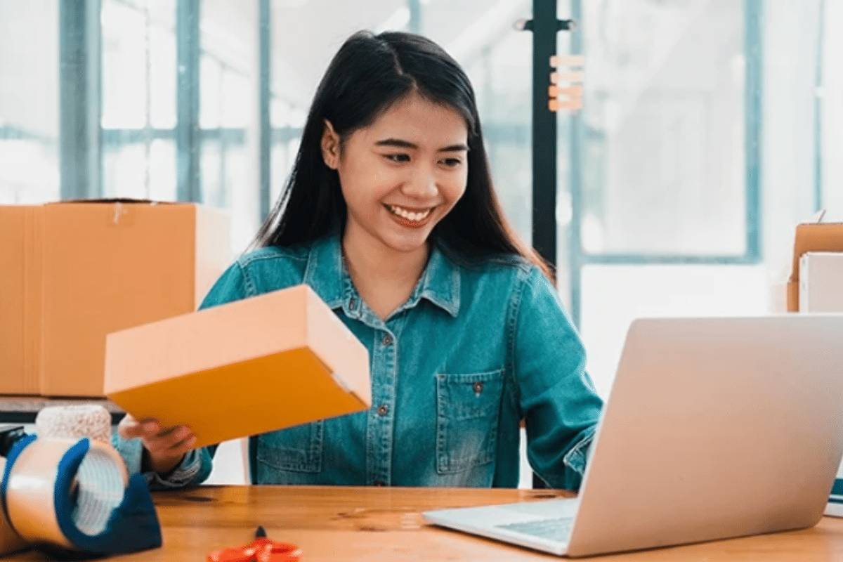 A woman joyfully holds a box and a laptop, radiating happiness.