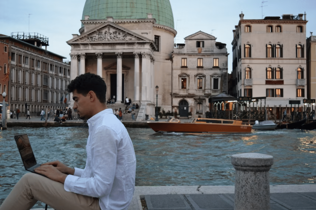 A man sitting on a bench by a canal, engrossed in his laptop, surrounded by serene scenery.